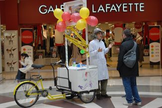 personne vêtue d'une blouse blanche qui explique quelque chose à un passant, à un stand dans un centre commercial